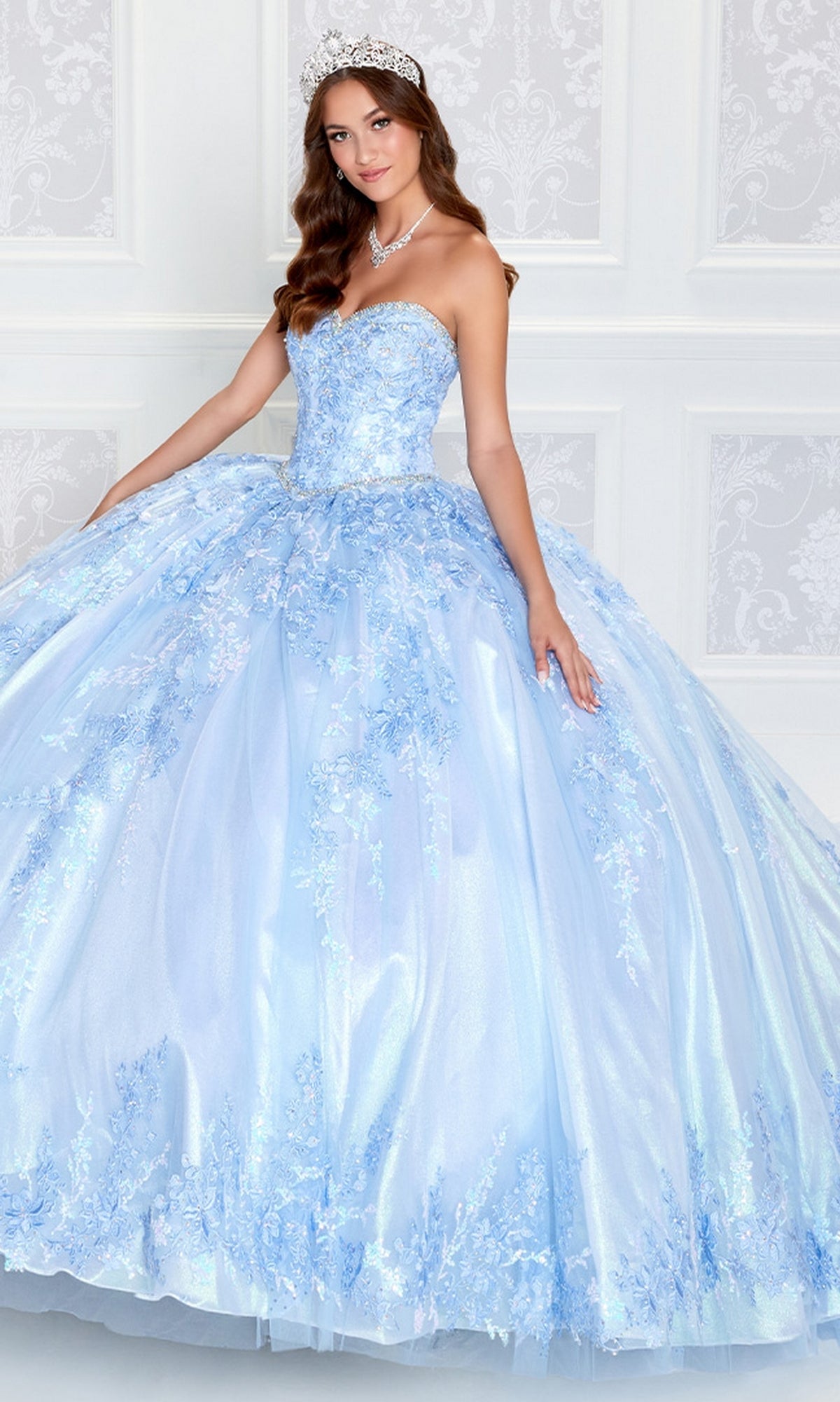 A woman wearing the Strapless Princesa PR12265 Quince Dress with Train, adorned with floral lace details and a tiara, stands in front of a white patterned wall.
